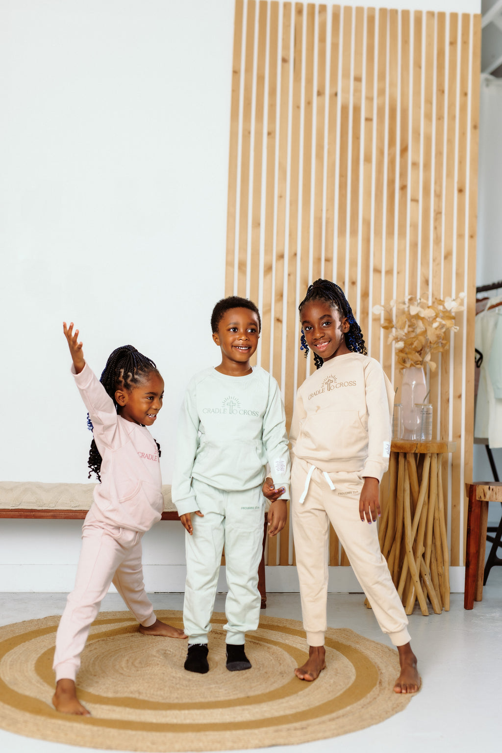 Three children posing together in a room with a wooden screen and round rug. Wearing crewneck with Cradle to Cross embroidered on the chest, a patch with Proverbs  22:6 on sleeve and Proverbs 22:6 embroidered on pant pocket 