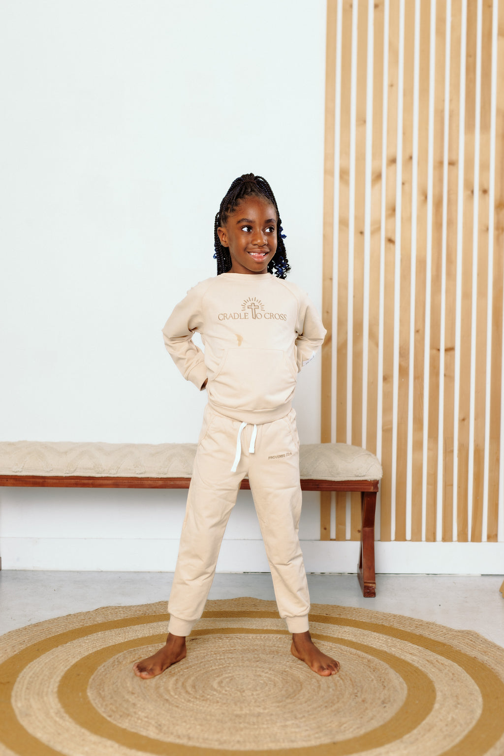 Young girl in beige sweatshirt and pants standing in a room with wooden bench and slatted wall.