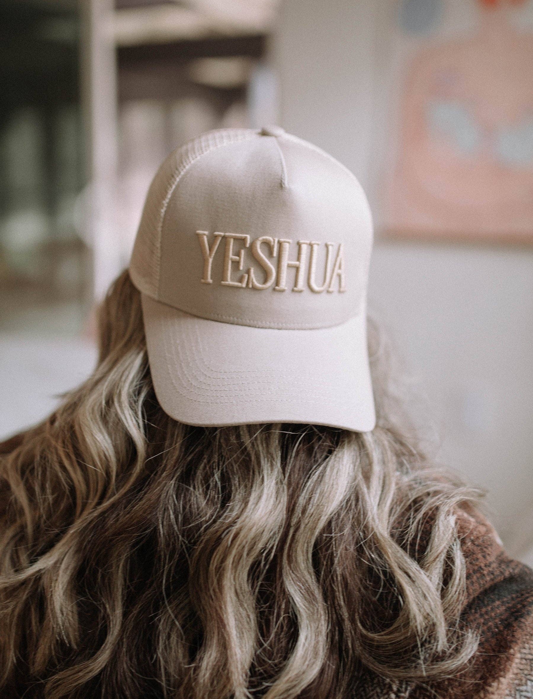 Beige cap with 'YESHUA' text worn by a person with wavy hair.